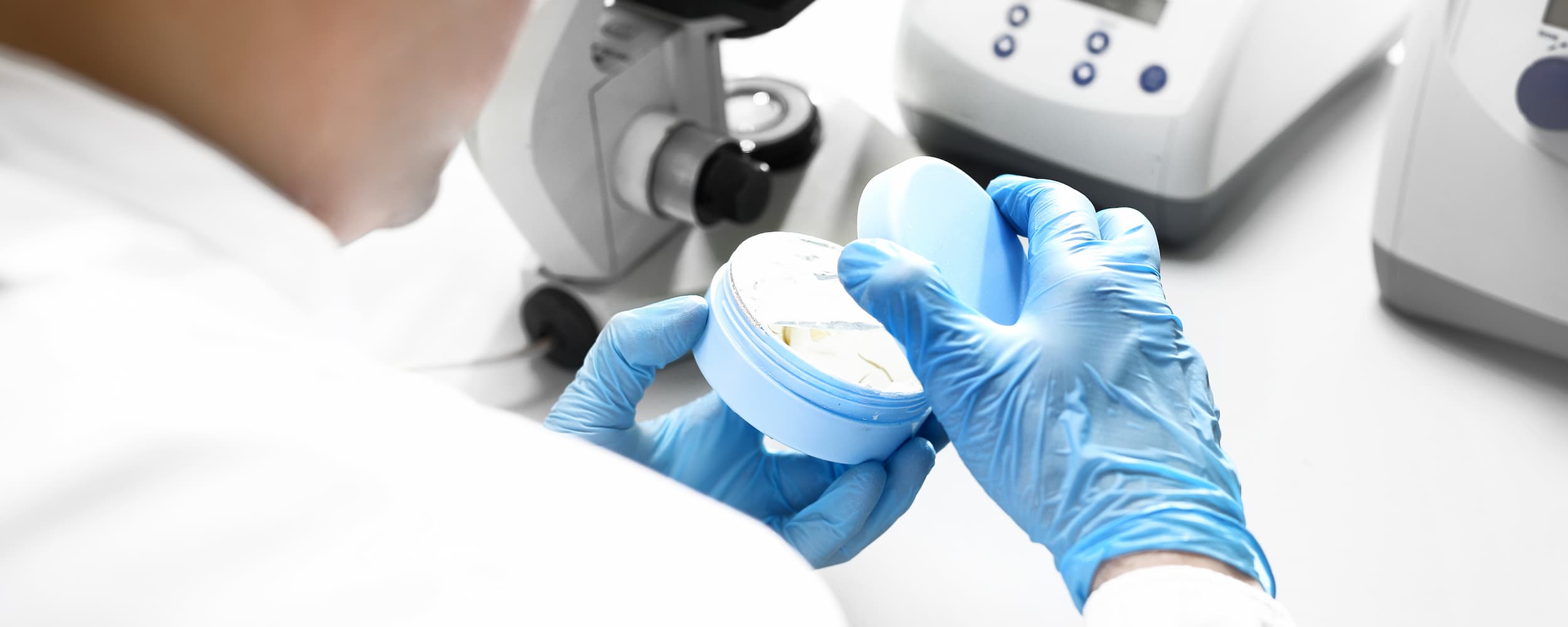 Person in a lab setting holding a small container with gloves on, surrounded by laboratory equipment.