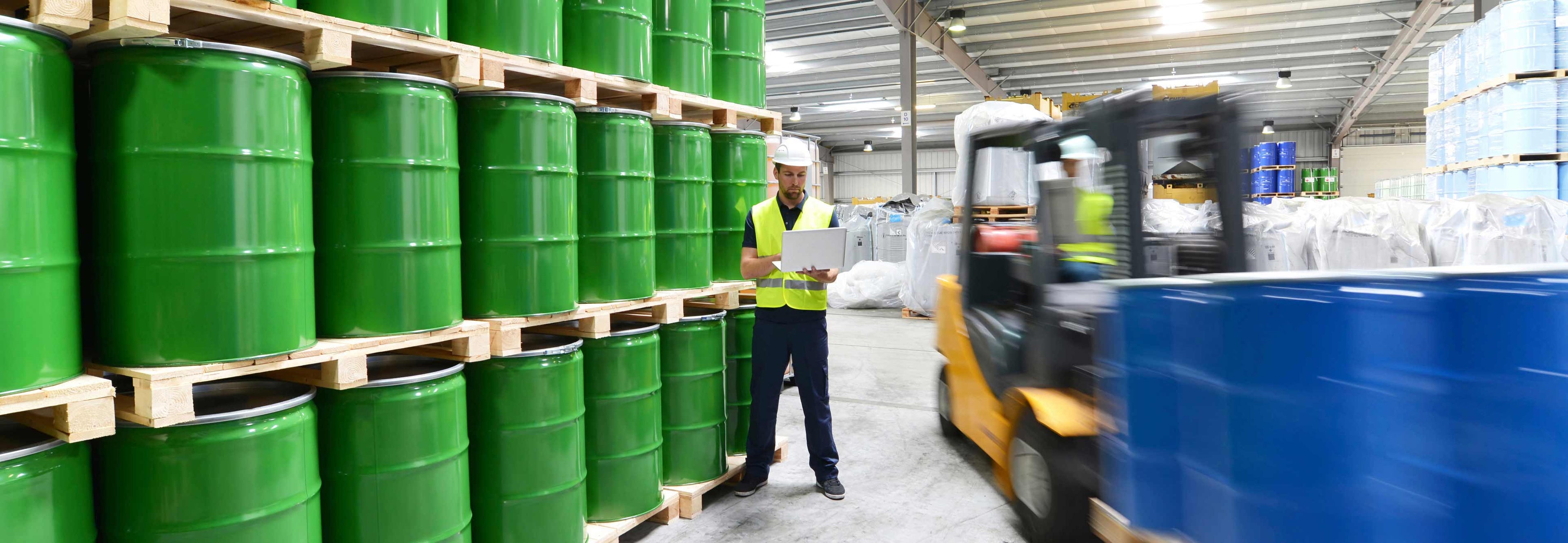Worker with a forklift in a warehouse with green barrels and blue containers.