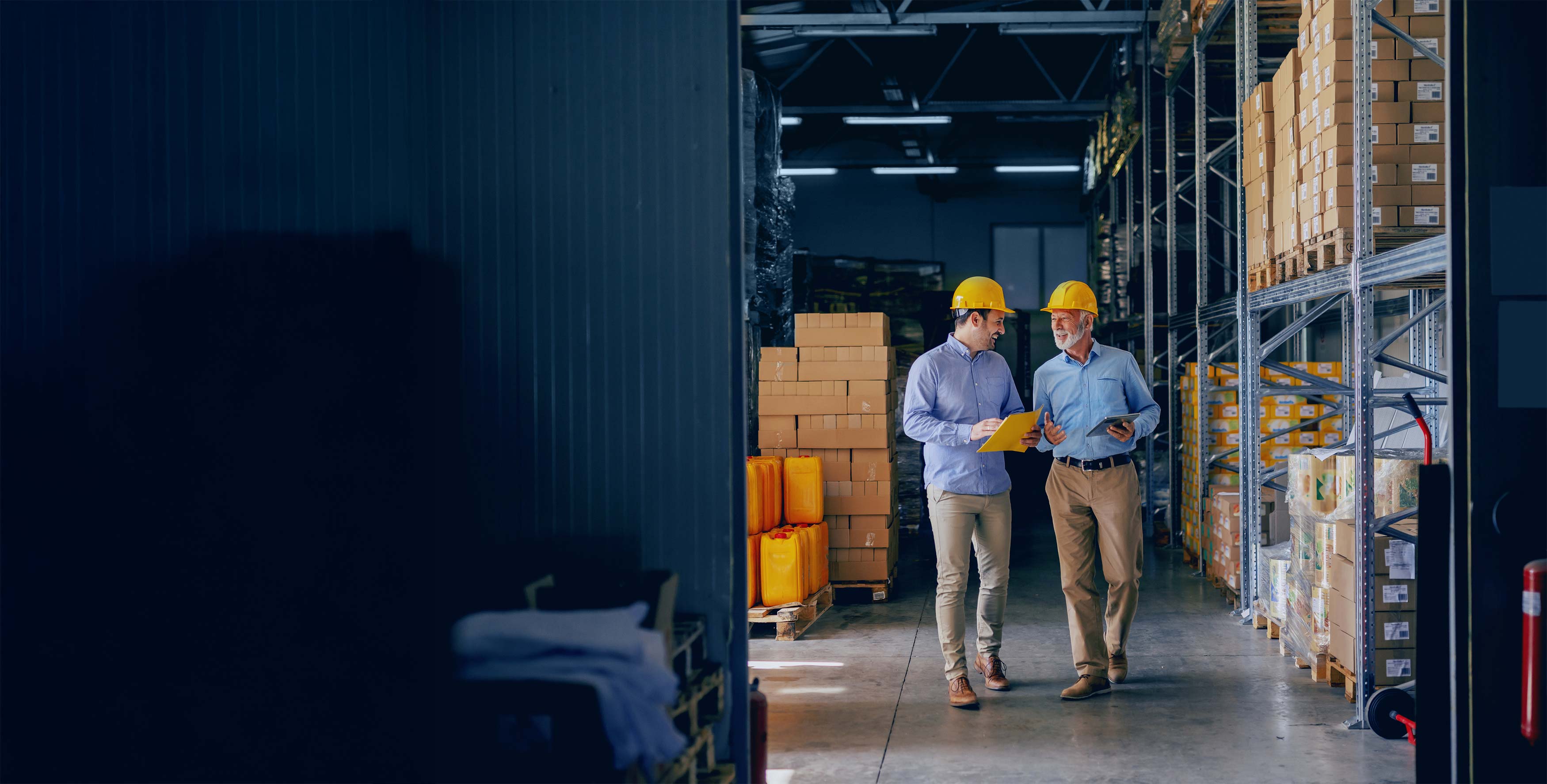 Two workers in a warehouse, one holding a tablet, the other walking, with shelves and boxes in the background.