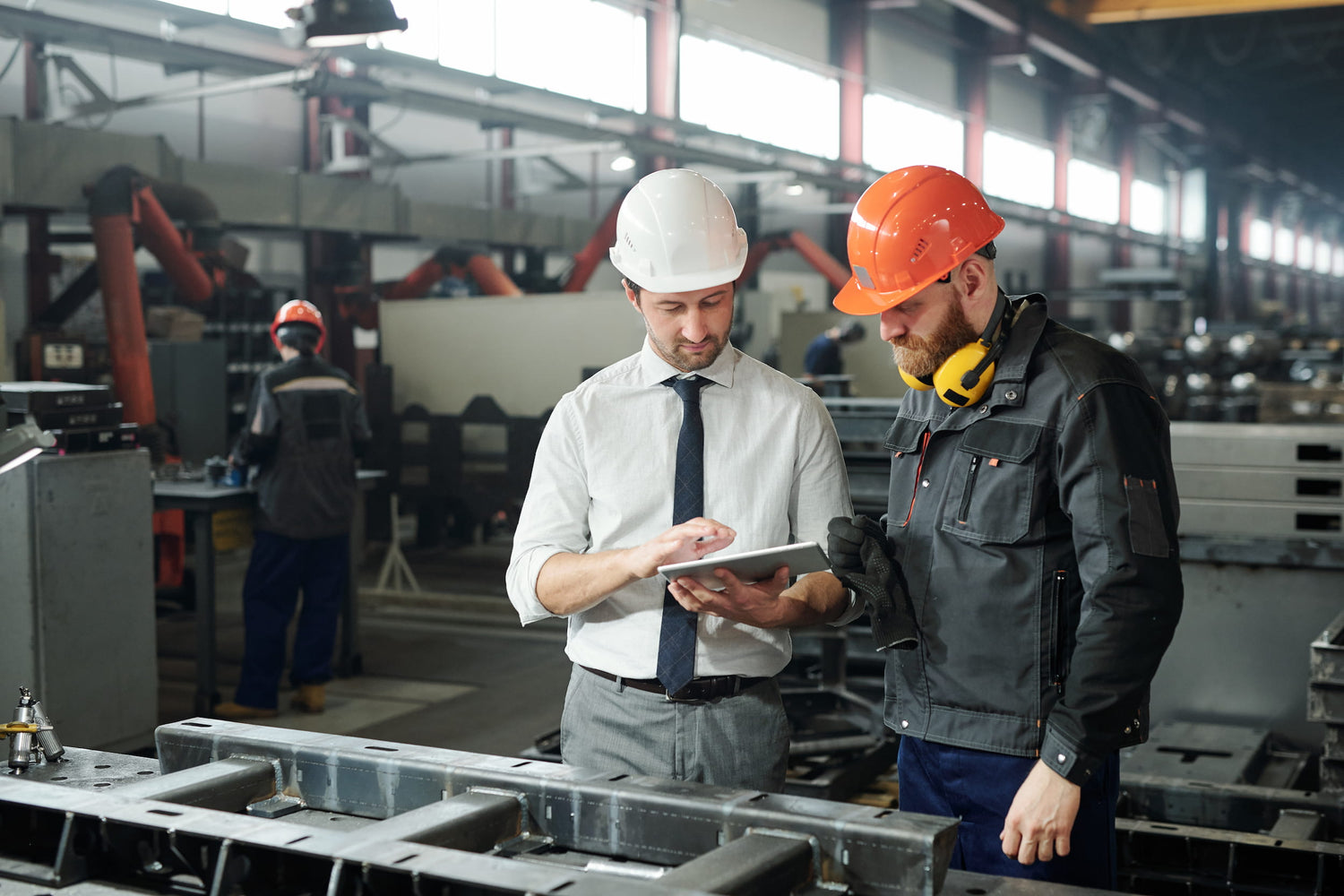 Two engineers in a factory setting, one holding a tablet, discussing work.