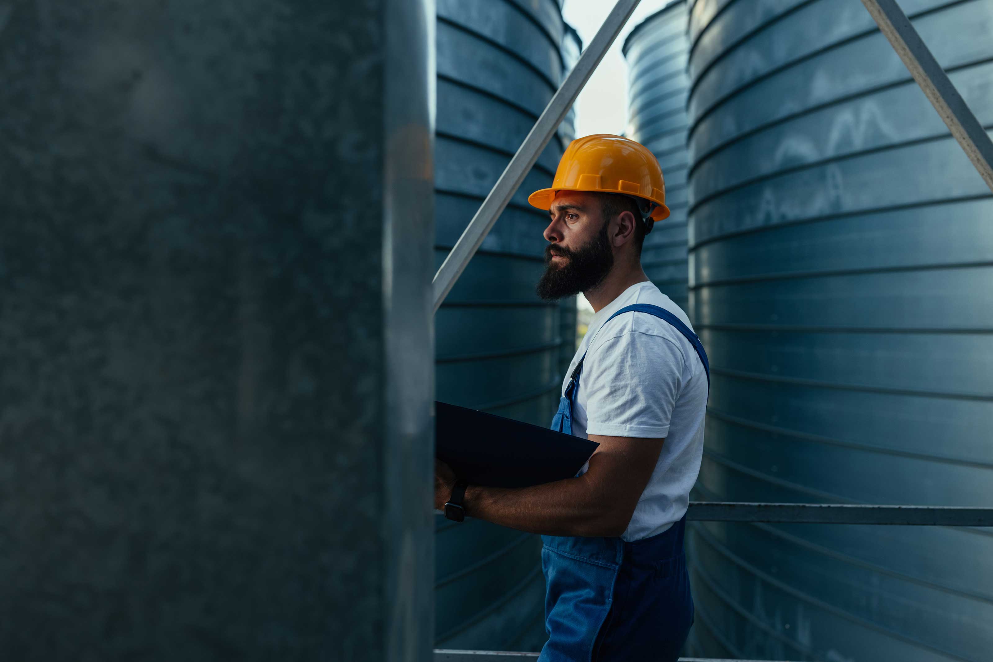 Man in a hard hat and overalls inspecting large cylindrical tanks.