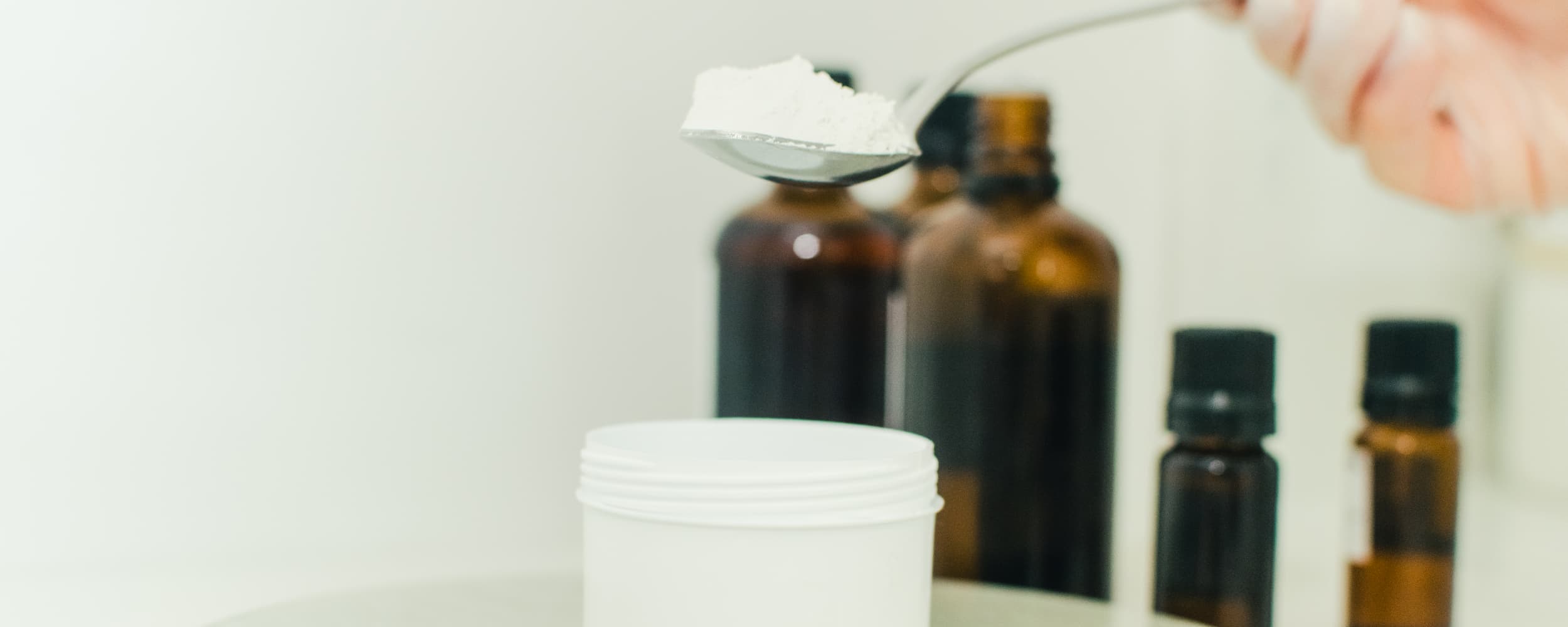 Person measuring a white powder with a spoon, surrounded by dark glass bottles on a light background