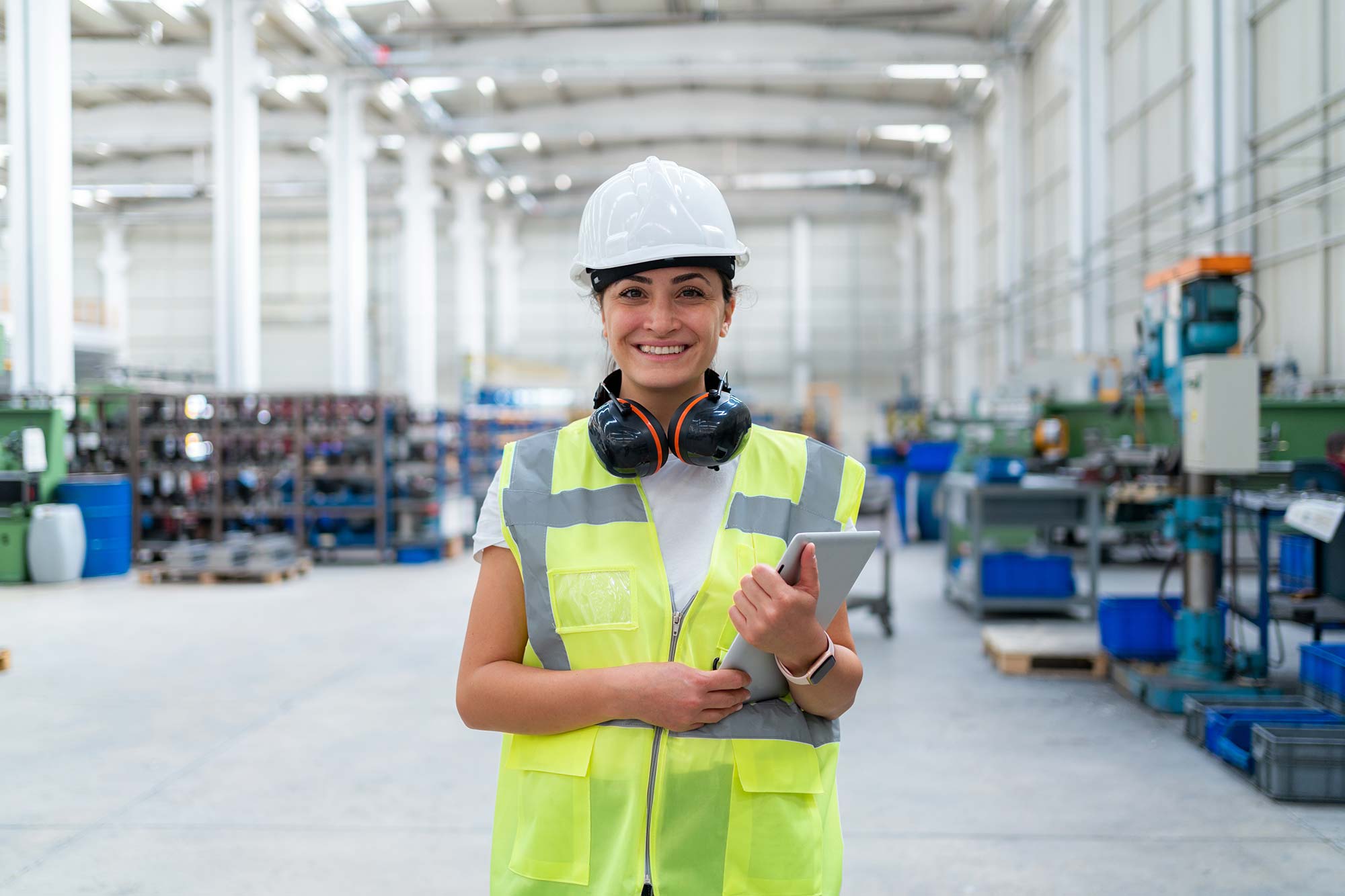 Person in a warehouse wearing a safety vest and hard hat, holding a tablet.