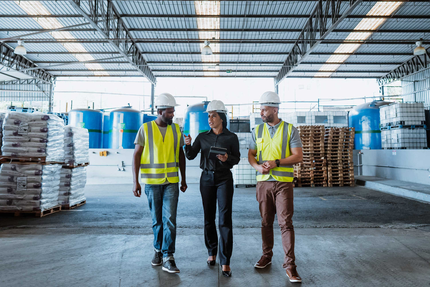 Three workers in a warehouse, two wearing safety vests and hard hats, with stacks of pallets and blue containers in the background.