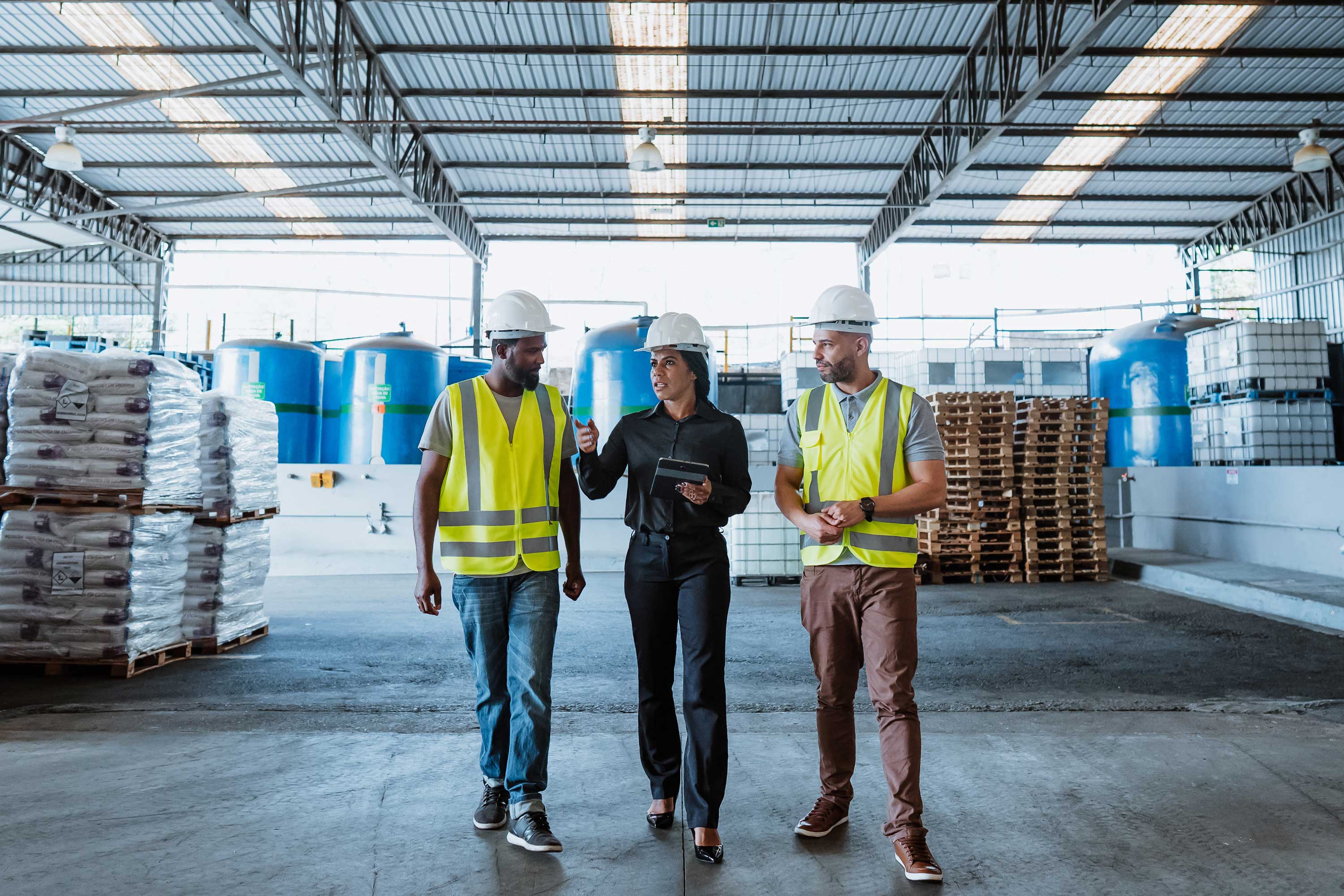 Three workers in a warehouse, two wearing safety vests and hard hats, with stacks of pallets and blue containers in the background.