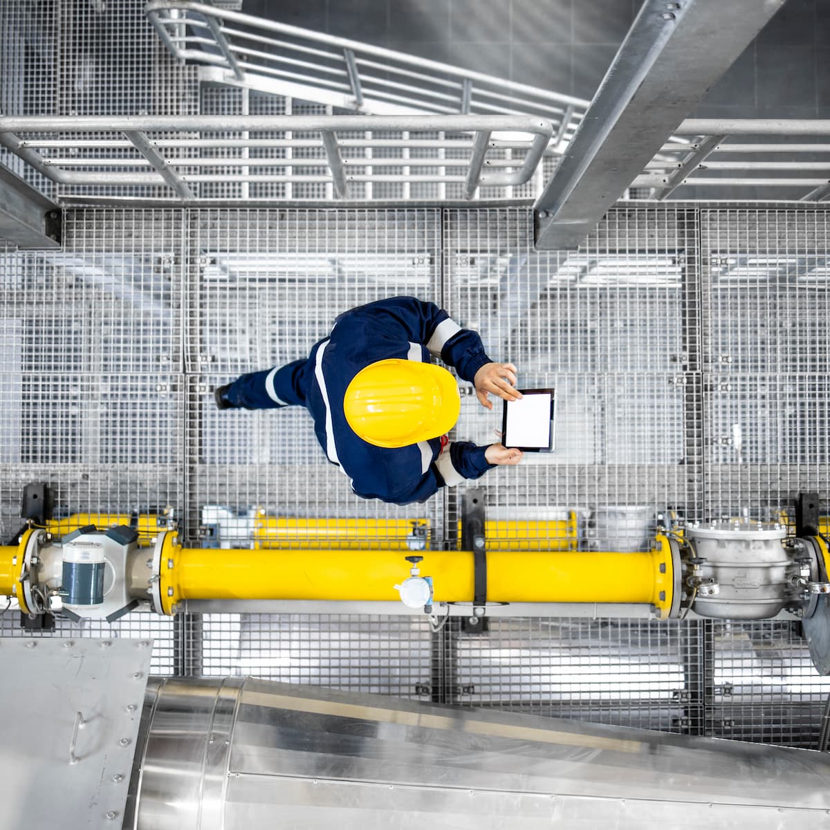 Person in a blue jacket and yellow hard hat using a tablet in an industrial setting with large pipes and metal structures.