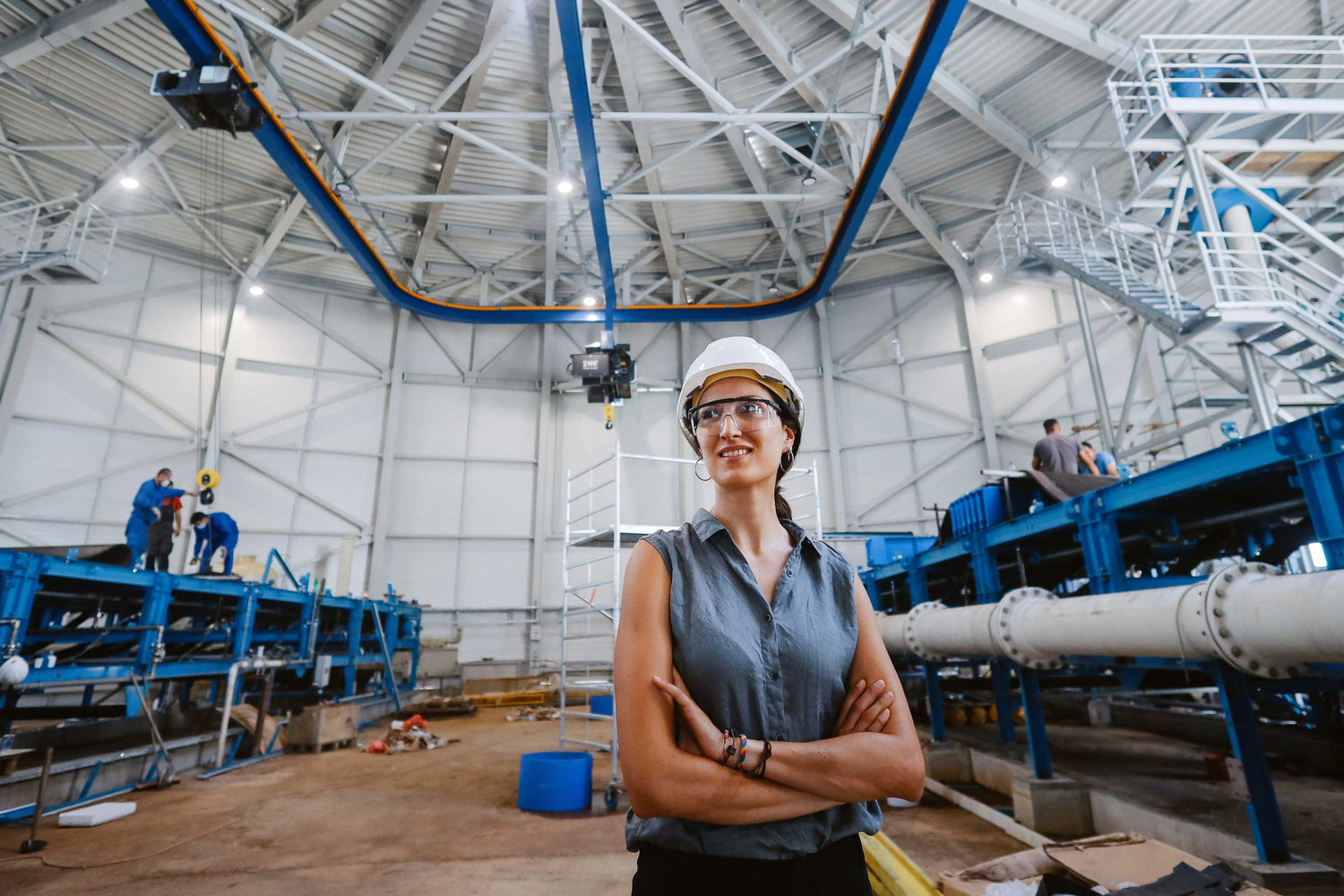 Person in a hard hat standing in a large industrial or warehouse setting with equipment and people in the background.