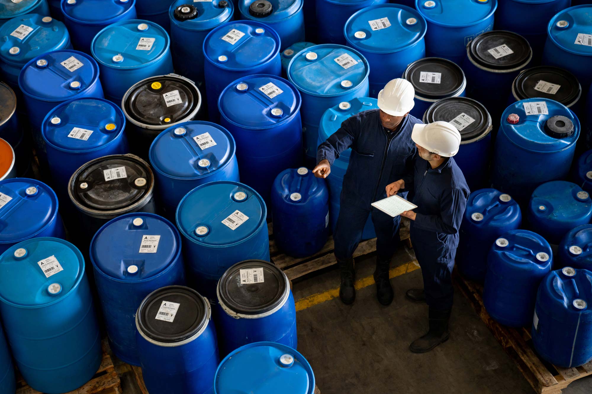 Two workers inspecting blue barrels in a warehouse setting