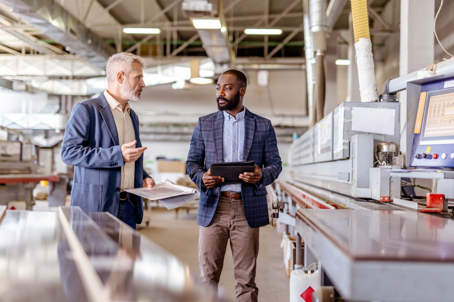 Two men in a factory setting, one holding a tablet and discussing with the other.