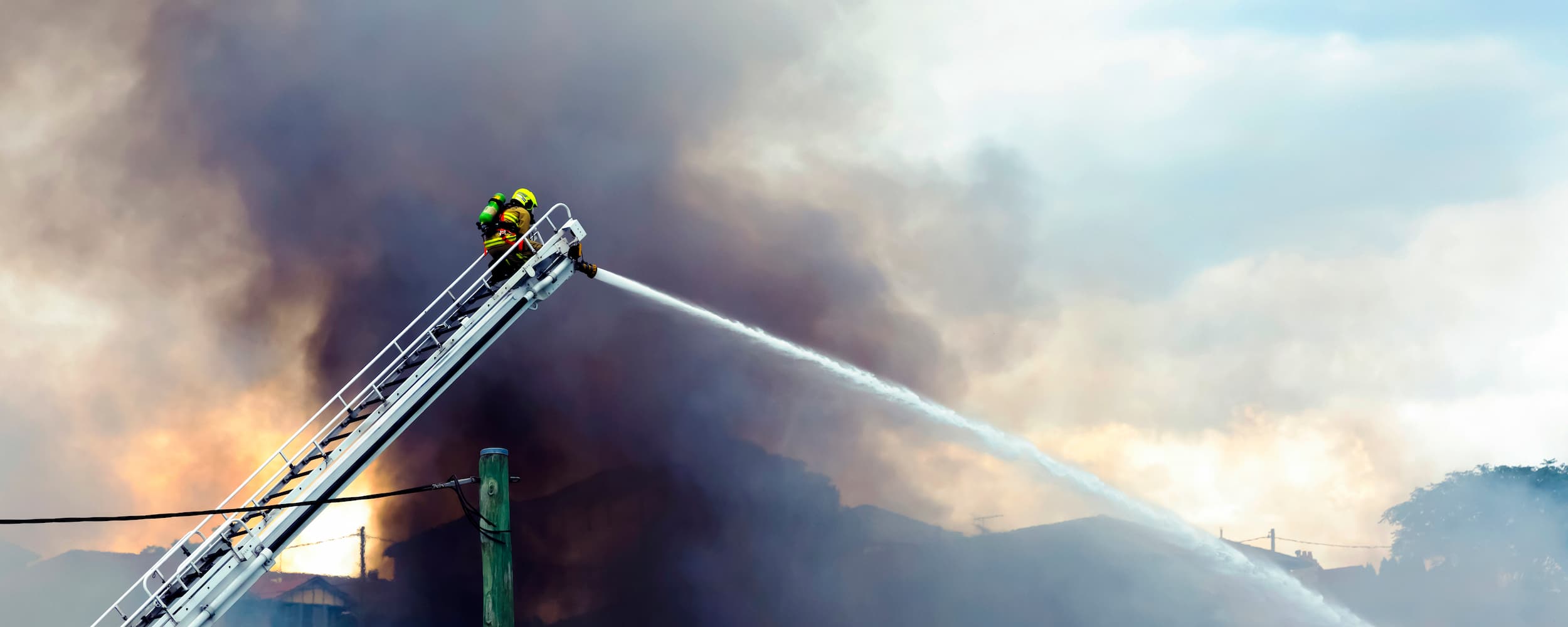 Firefighter on a ladder with hose combating a fire against a dark sky.