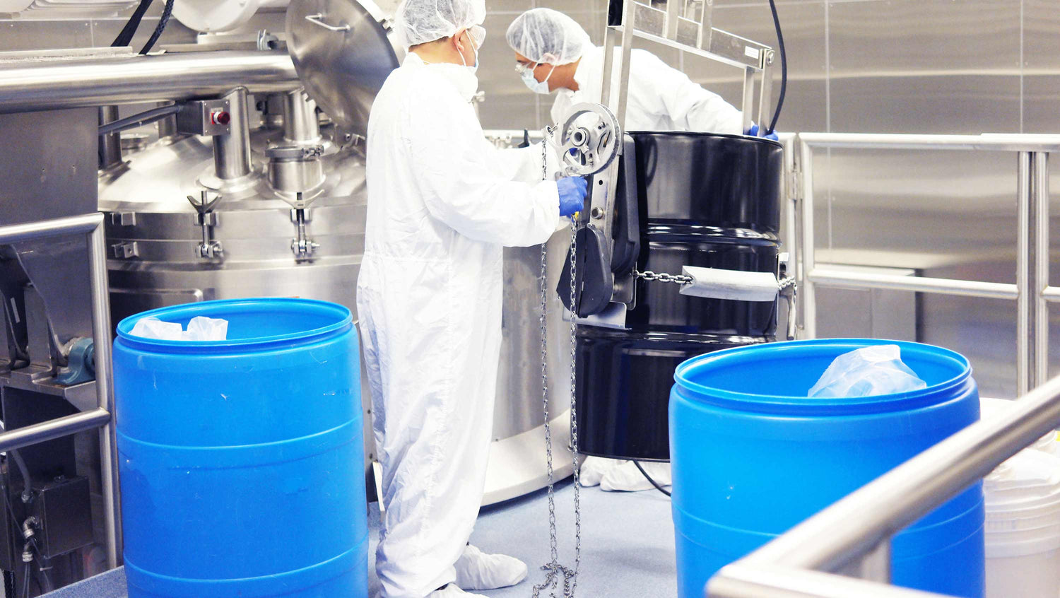 Workers in a sterile environment with blue barrels and industrial equipment.