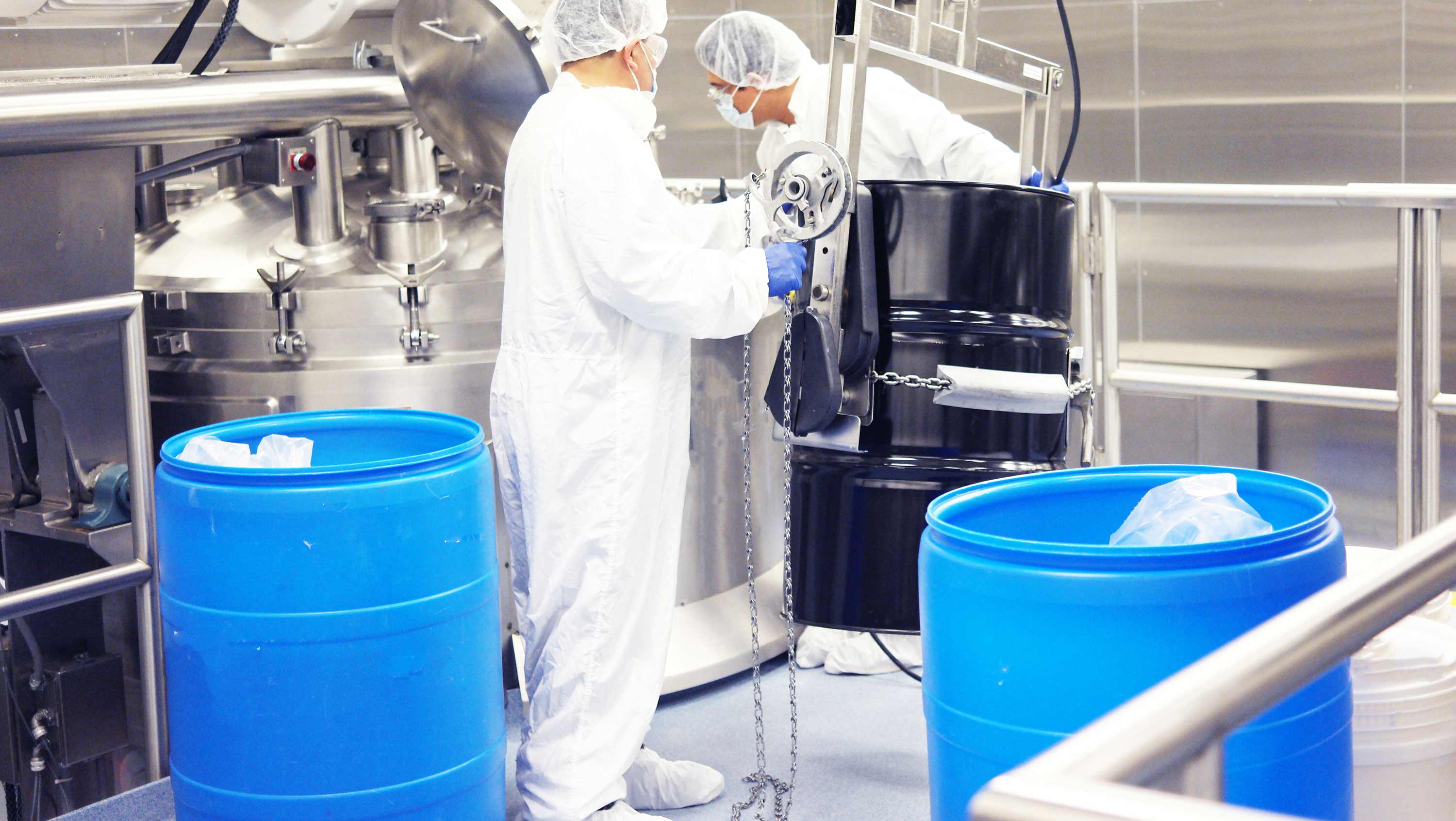 Workers in a sterile environment with blue barrels and industrial equipment.