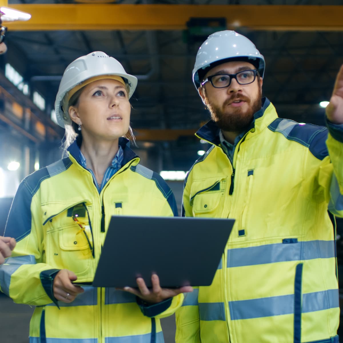 Two engineers in high-visibility jackets and hard hats examining a laptop in an industrial setting.