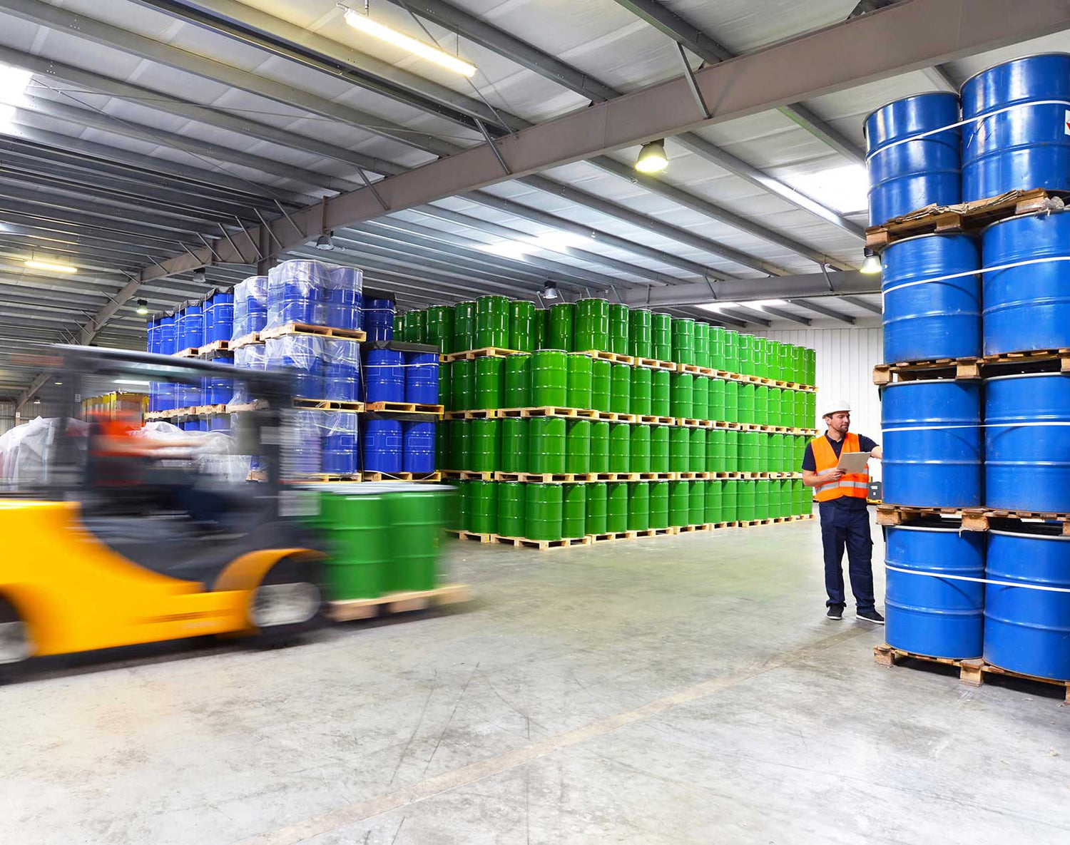 Warehouse interior with blue and green barrels on pallets, a forklift, and a worker.
